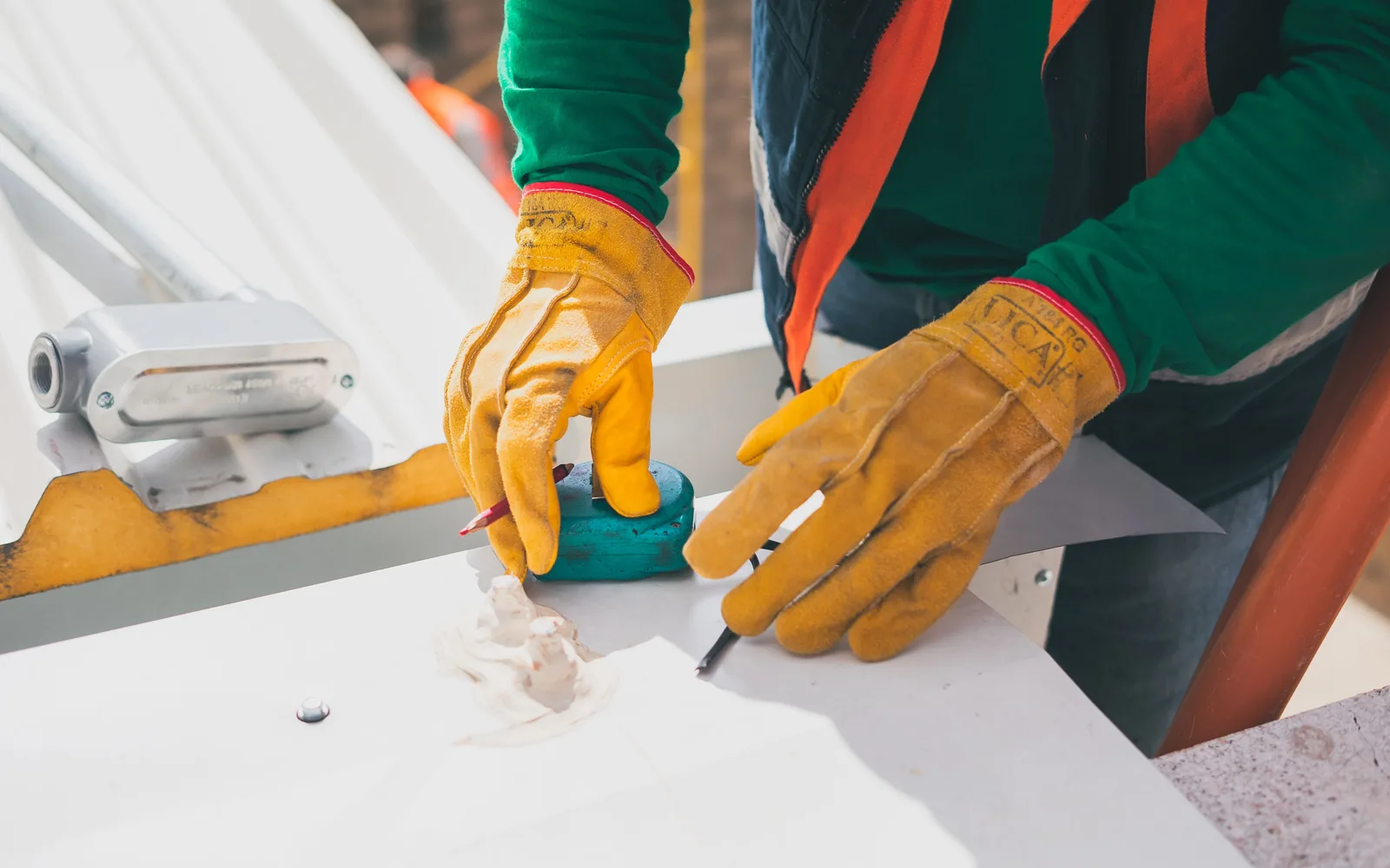 Worker measuring and marking a metal roof or flashing component while wearing protective gloves.
